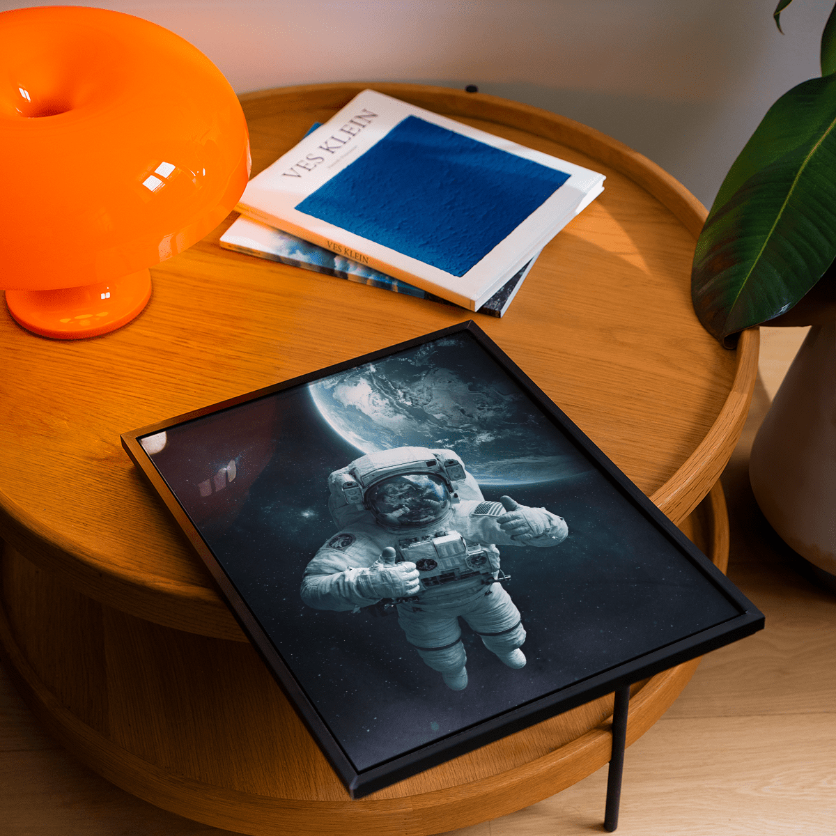 Astronaut wall art in a black frame on a wooden side table with a bright orange lamp