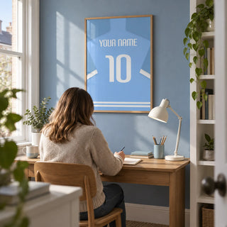 Football Shirt Poster in Light Blue with White Sleeve Stripes, framed on the wall of a compact home study in a UK terrace with a muted blue wall and white shelving
