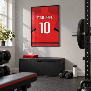 Football Shirt Poster in Red with Black Sleeve Bands, framed on the wall of a tidy converted spare-room home gym in a UK house with a pale grey wall and plain dark rubber floor