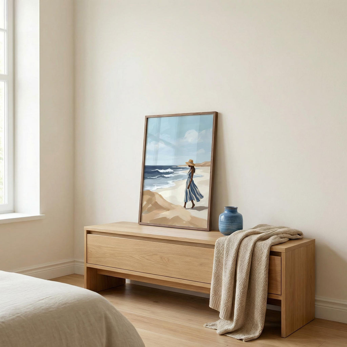 Beach Woman Art Print leaning on a wood sideboard with a blue vase and cream throw beside a bright window