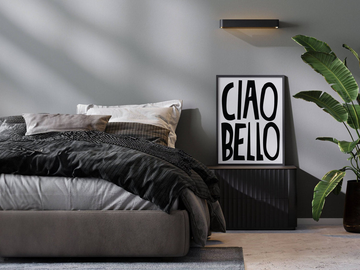 Italian wall art with ciao bello text in black and white resting on a bedside table with a green plant nearby