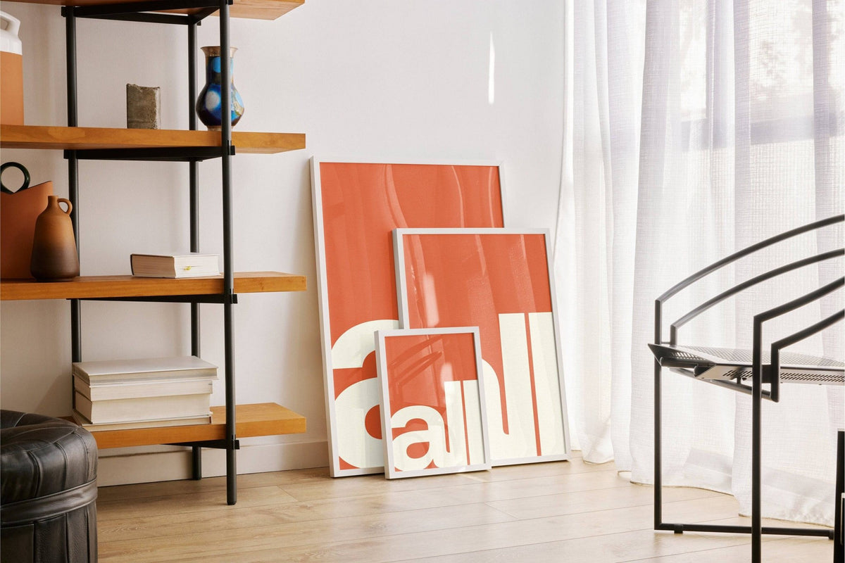 Modern Typography Art prints with letters al in cream and orange leaning on a floor near a shelving unit and chair