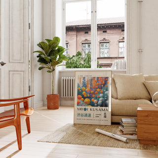 Abstract floral wall art with round pom-pom flowers in orange and blue leaning by a beige sofa near a green plant