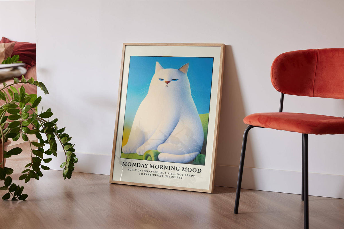 Monday Mood Cat Poster leaning on a floor beside a green plant and a rust red chair