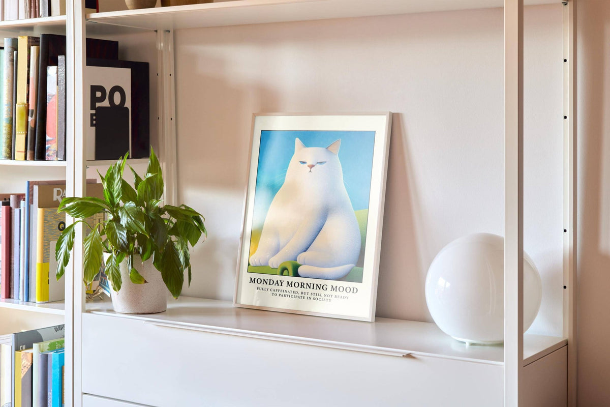 Print of Monday Mood Cat Poster with white cat sitting framed in white leaning on a shelf next to a green plant and a white round lamp