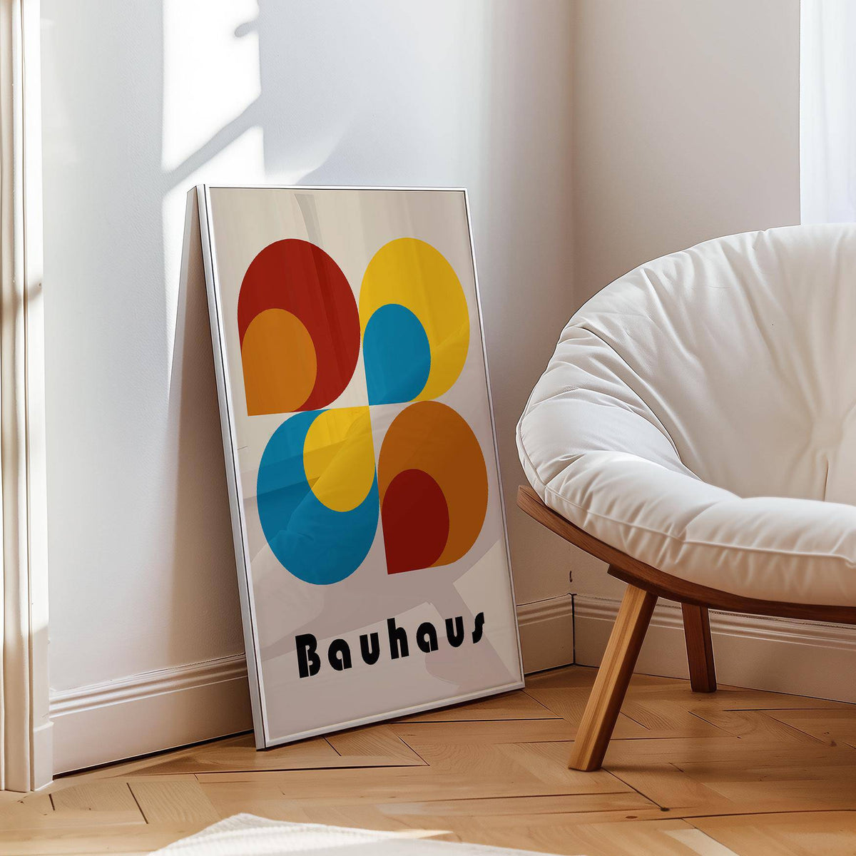 Print leaning on floor of geometric prints with colourful bauhaus shapes beside a white cushioned chair