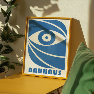 Framed print Bauhaus with stylised blue eye and text leaning on a yellow table by green cushion