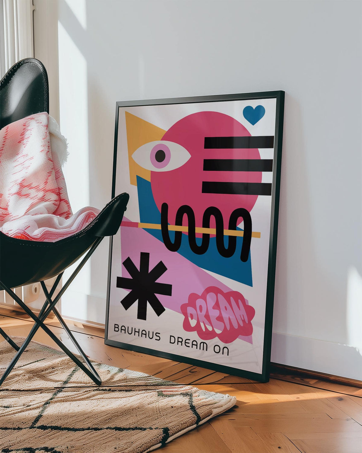 Maximalist wall art with abstract eye and shapes in pink yellow and blue leaning against a white wall by a chair