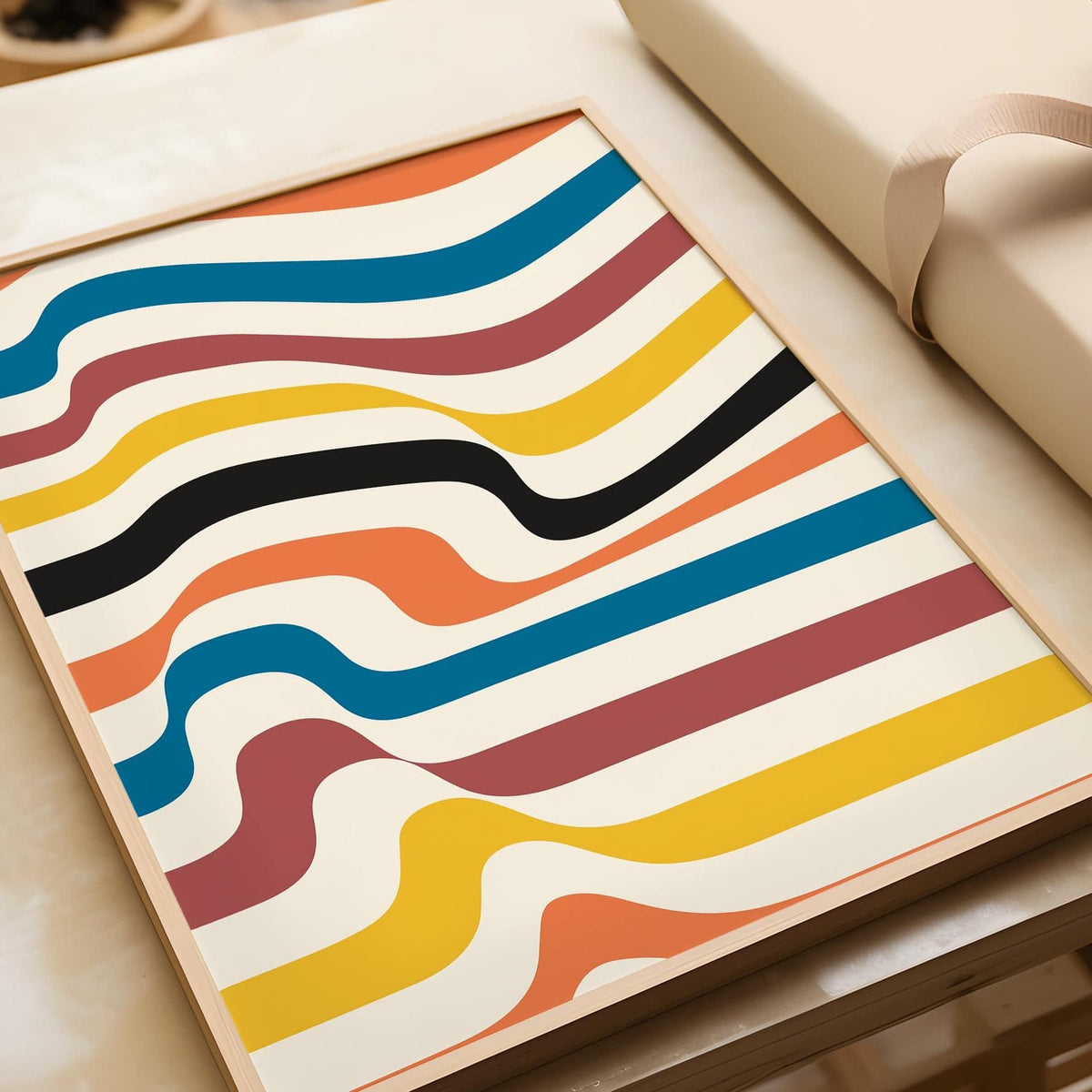 Framed close-up of Art Work for Wall with wavy colourful stripes in blue, orange, black and yellow on a table near beige wrapping paper