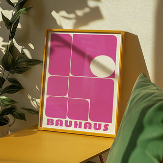 Framed wall art pink with bauhaus geometric shapes on a yellow surface near a green cushion and plant