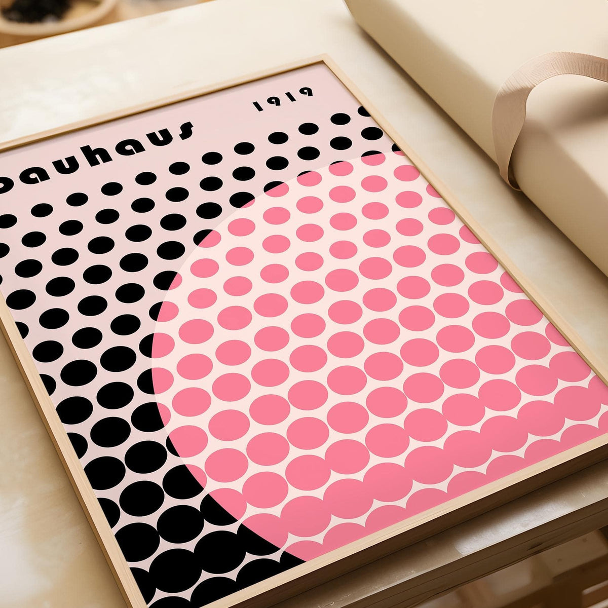 Framed Bauhaus print poster with pink circle and black dots leaning on a table with a ribboned package
