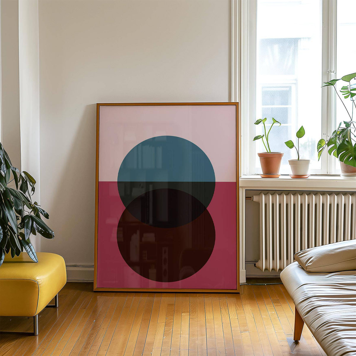 Print leaning on a wooden floor of abstract overlapping circles in blue and brown with a pink background beside houseplants