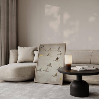 Print leaning on the floor of flying geese in beige and grey near a black round table and lamp