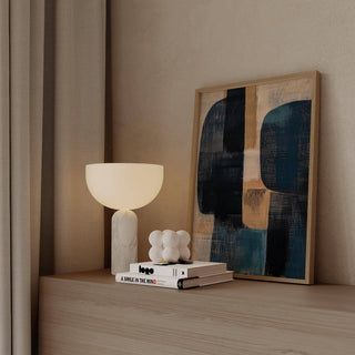 Print leaning on a shelf with a beige base and navy and tan abstract shapes with a white lamp nearby
