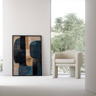 Print on a wall in a bright room showing abstract shapes in navy blue and beige next to a cream modern armchair