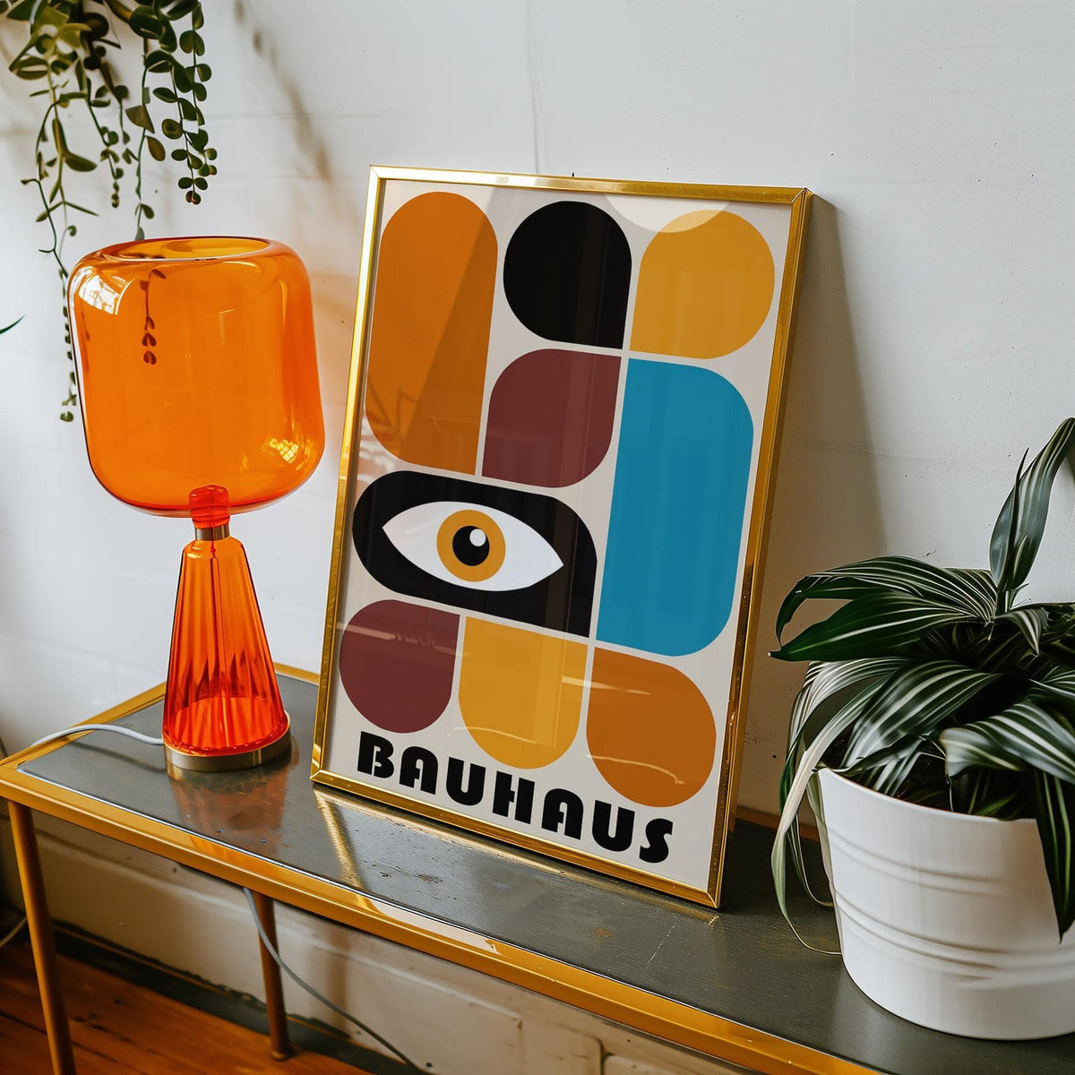Framed print of geometric shapes wall art with abstract eye in warm and blue tones on a table with orange lamp and white plant pot