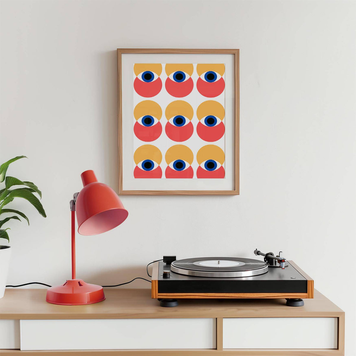 Wall Art Eye framed print with abstract eyes pattern in red and yellow near a red desk lamp on a wooden sideboard