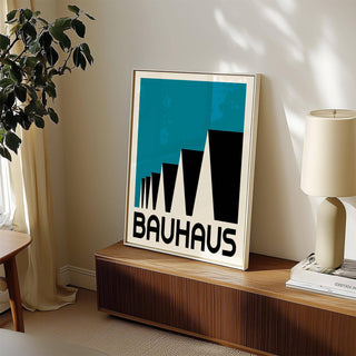 Print leaning on a shelf of Bauhaus poster blue with abstract stepped rooftops and bold typography beside a cream lamp