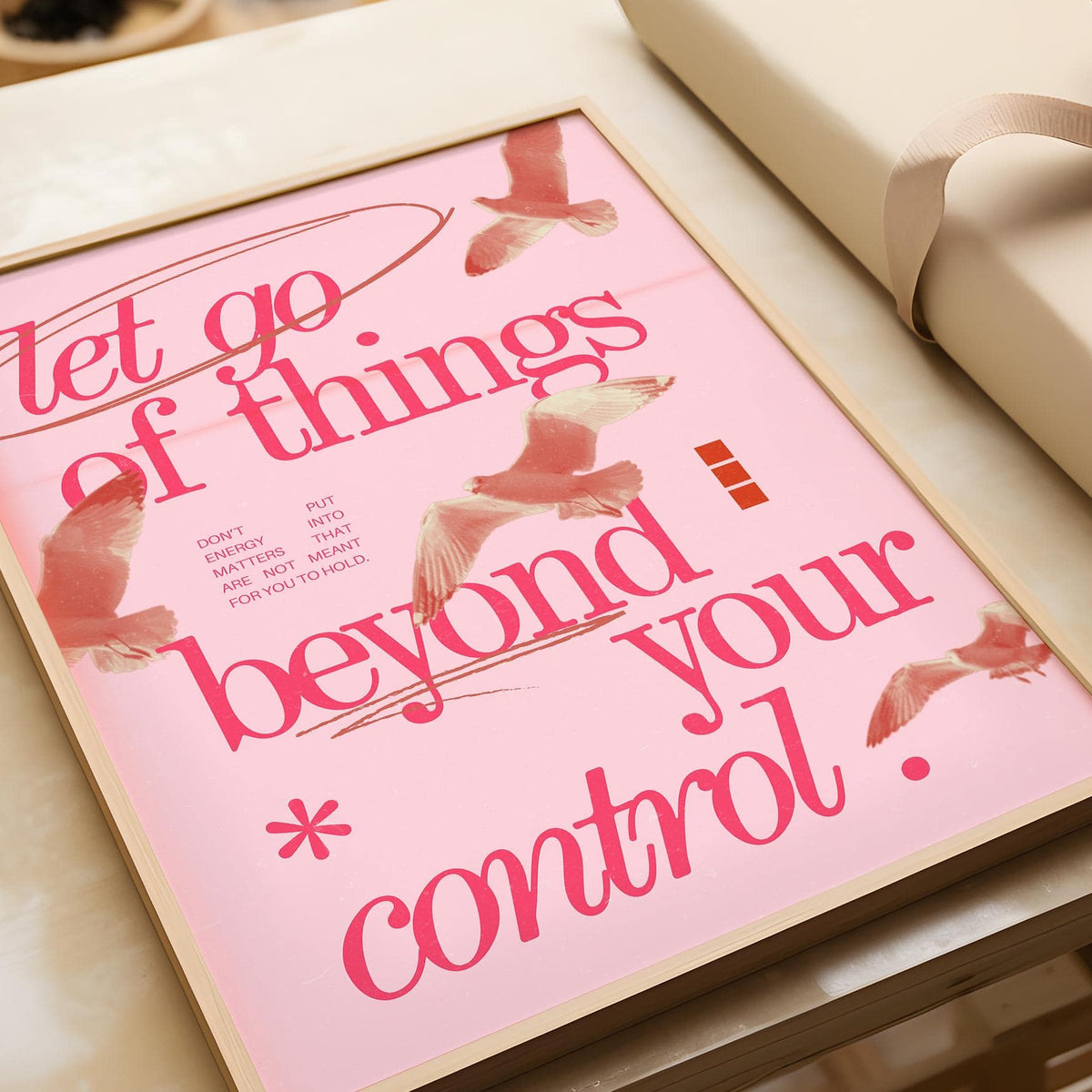 Framed pink and red Let Go Typography print leaning on a wooden surface next to a beige gift box