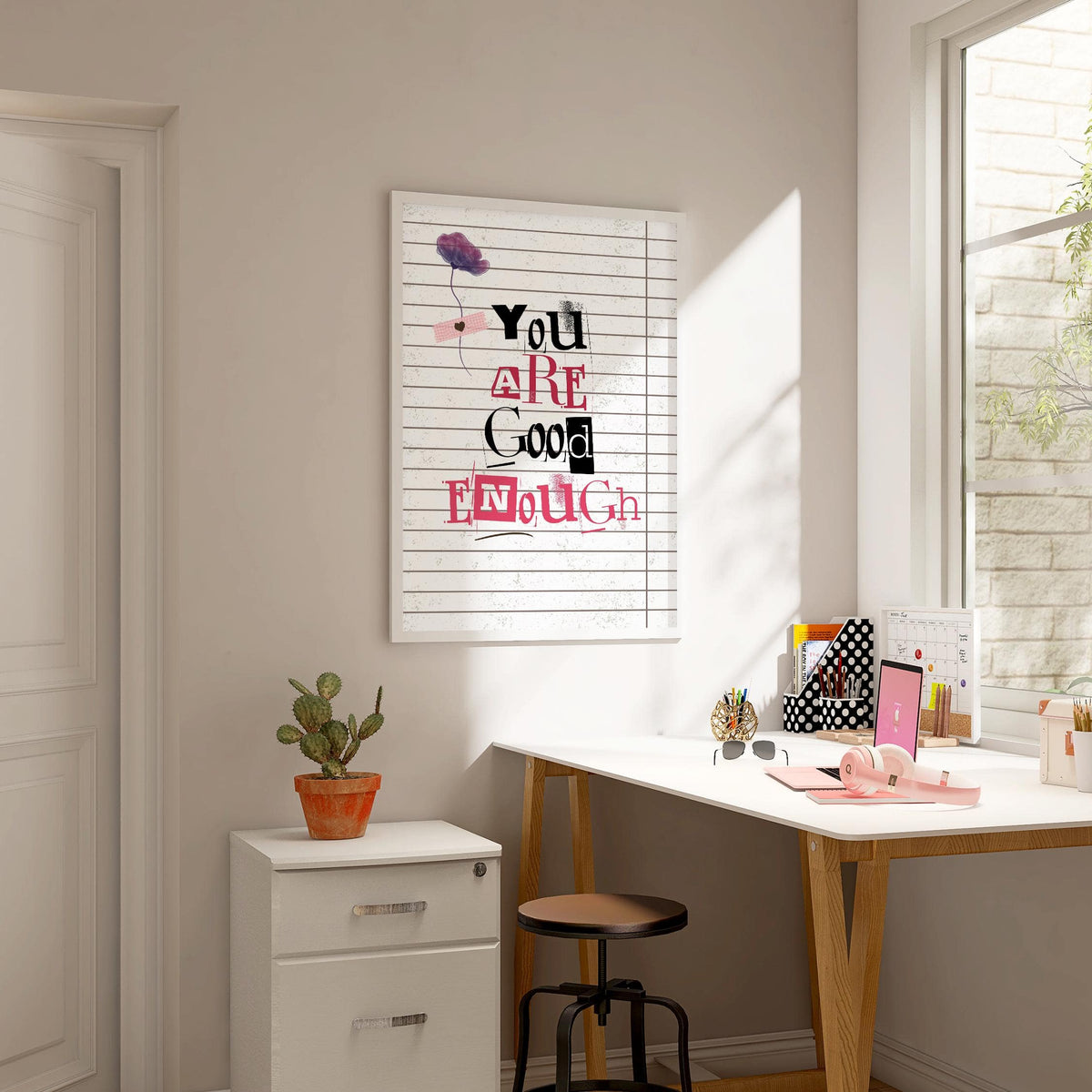 Posters of Positivity wall art with mixed pink and black text above a green potted cactus by a white desk in a light office