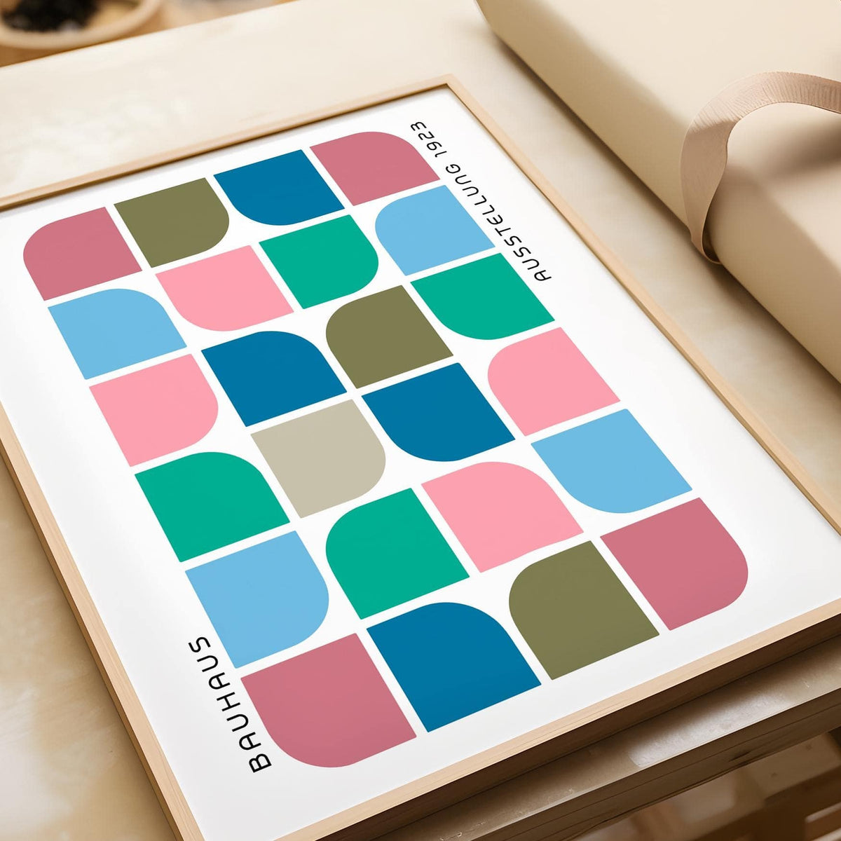 Detail close-up of Block Colour wall art with geometric block shapes in pink blue olive and teal on a table near wrapping paper