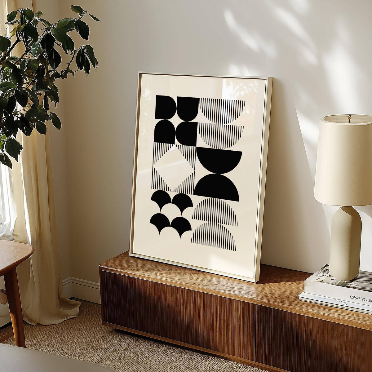 Print leaning on a cabinet of black and white geometric wall art in a neutral room with a lamp and plant