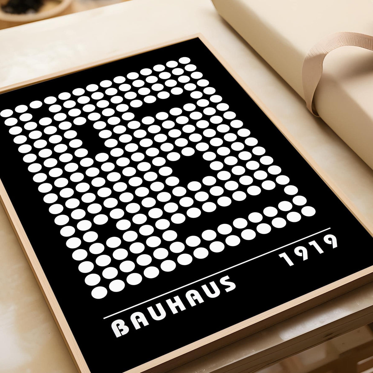 Framed Digital Wall Art with white dots in a bauhaus style on a black background leaning on a beige surface near a wrapped gift box