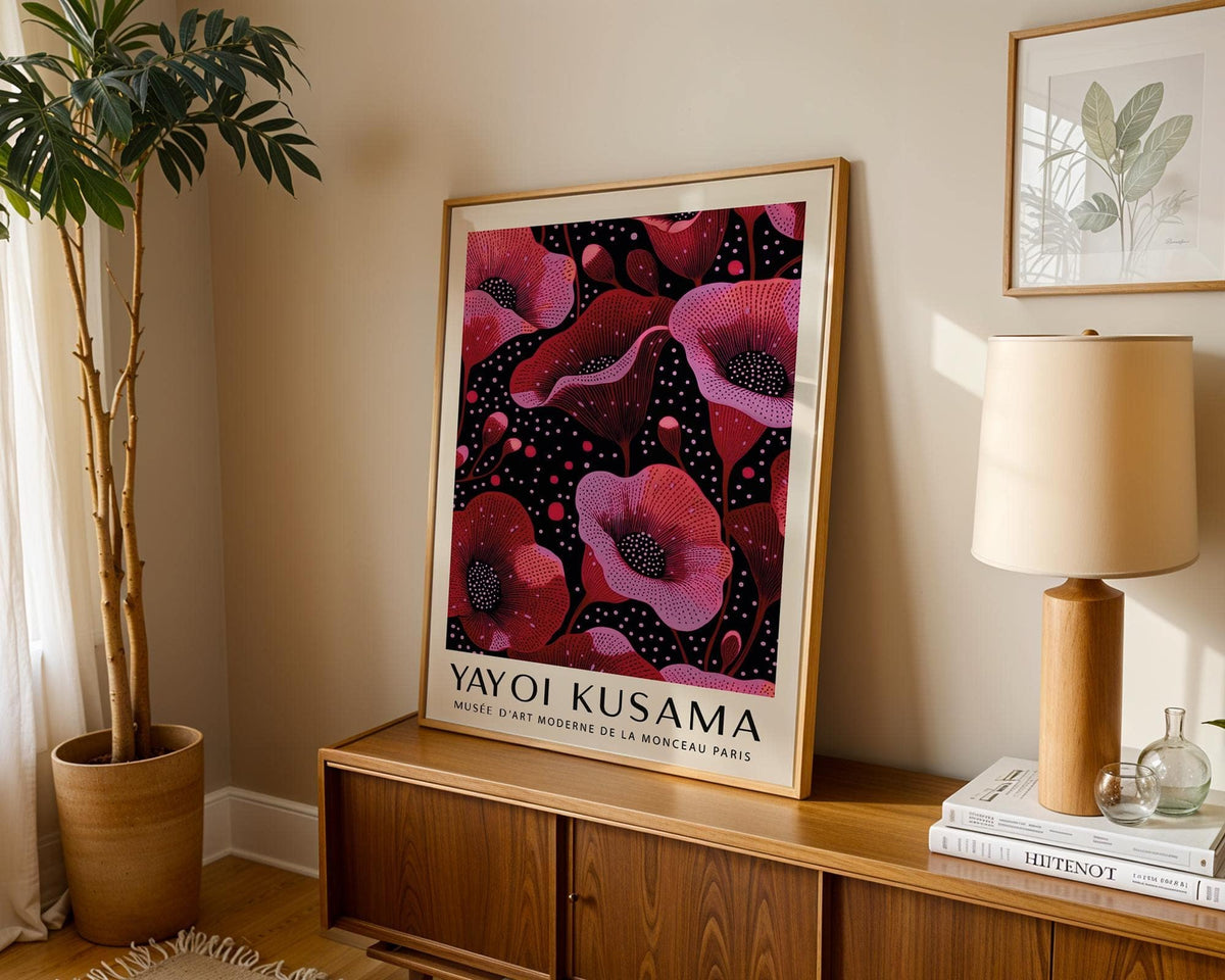 Print leaning on a wooden sideboard with stylised pink poppies and black background beside a lamp and plant