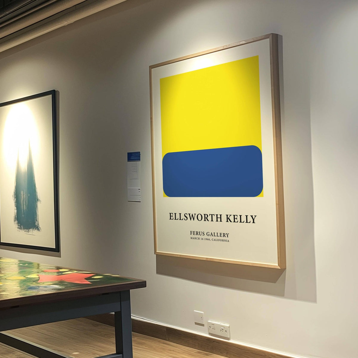 Minimalist artwork with blue rounded rectangle on yellow square in a gallery with table nearby