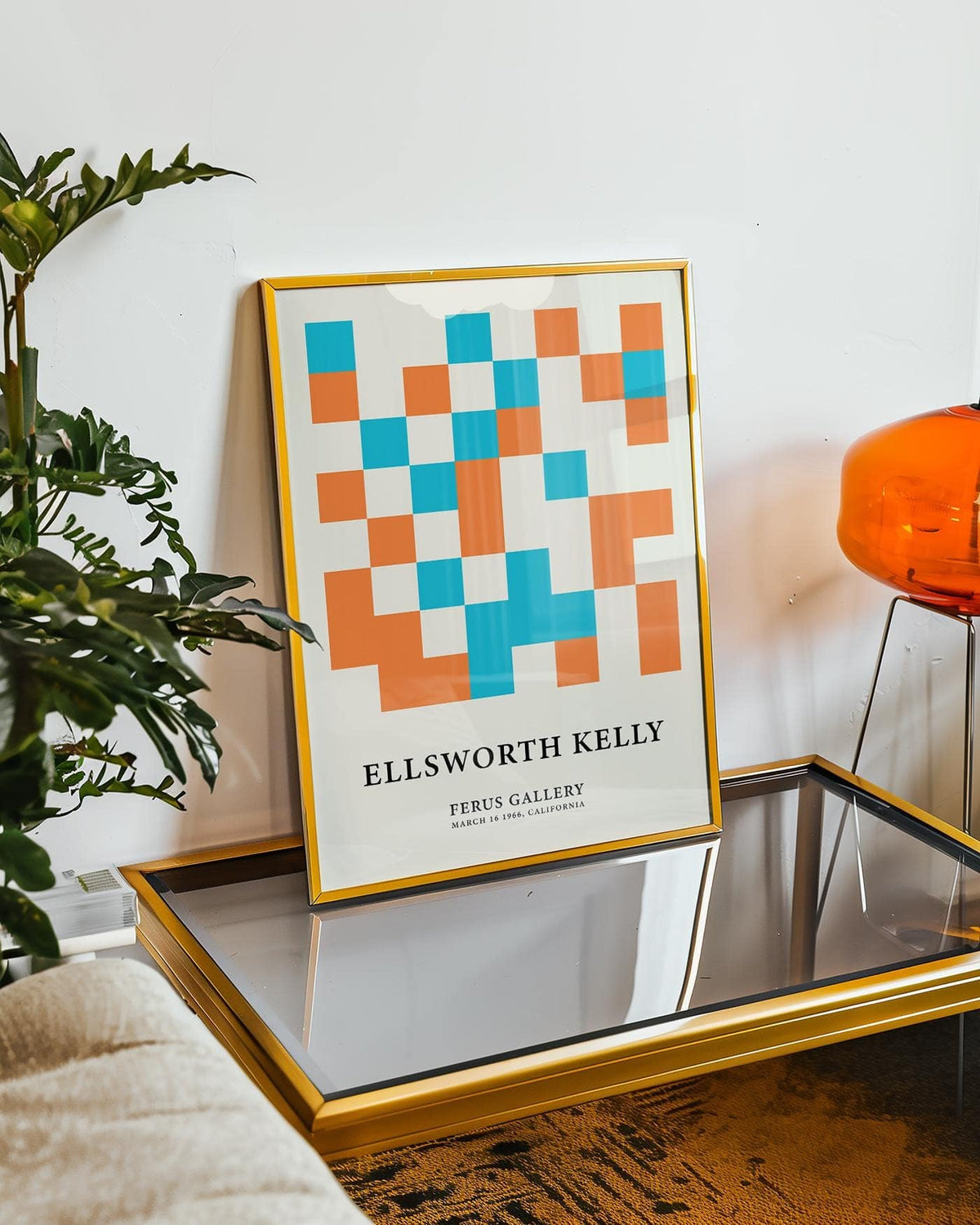Print leaning on a glass table with orange and blue squares and golden frame near a plant