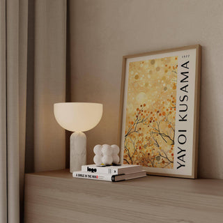 Print leaning on a shelf with autumn branches and yellow dots beside a lamp and books