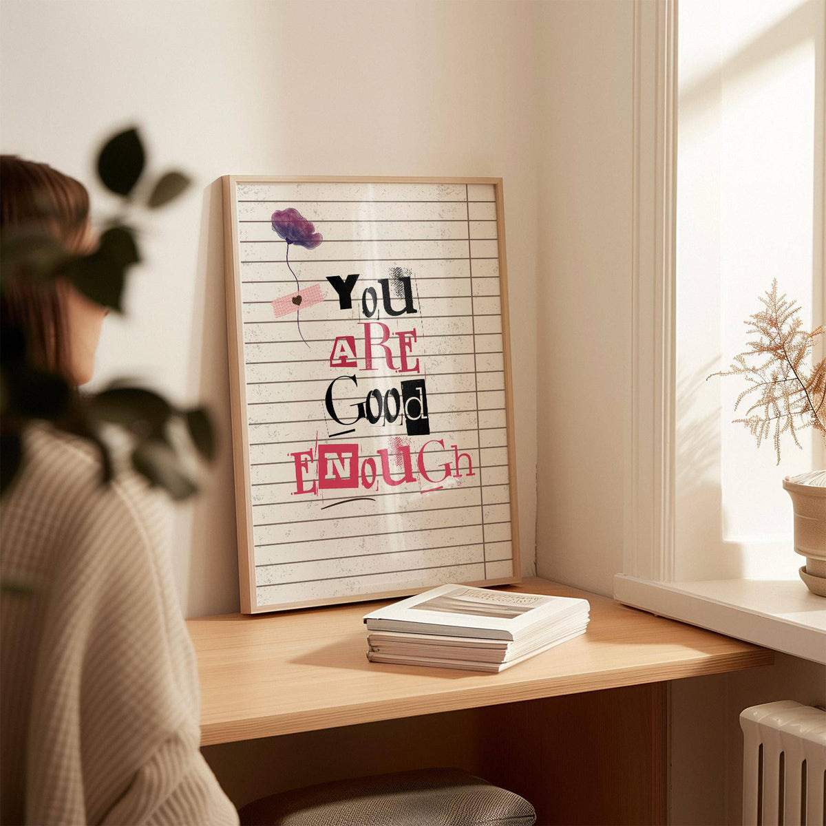 Posters of Positivity leaning on a desk with a stack of books and a potted plant nearby in soft natural light