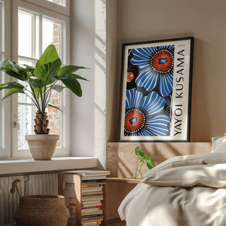 Print of blue poppy flowers with red and black highlights leaning on a bed headboard beside a plant and books