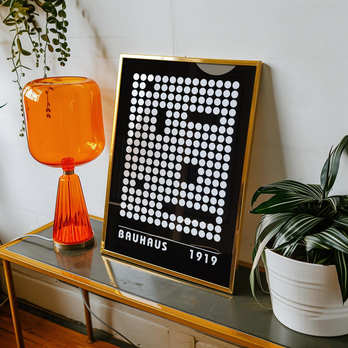 Print leaning on a shelf showing abstract dot pattern with white dots on black with gold frame next to orange lamp and green plant