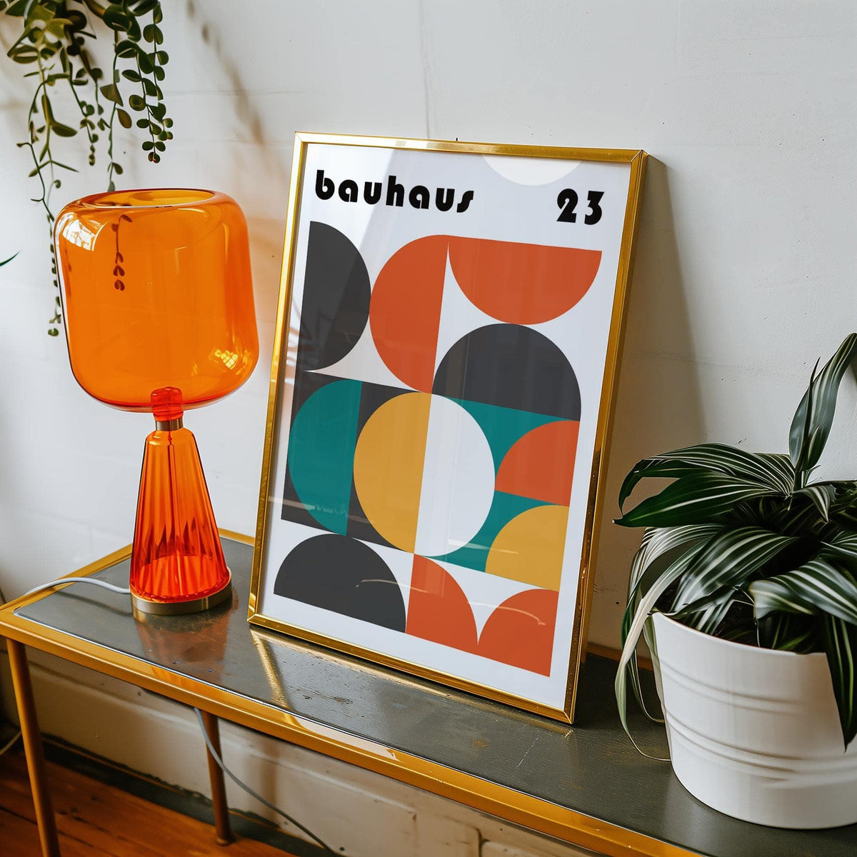 Framed MCM wall art with geometric shapes in orange black and teal leaning on a glass table with an orange lamp