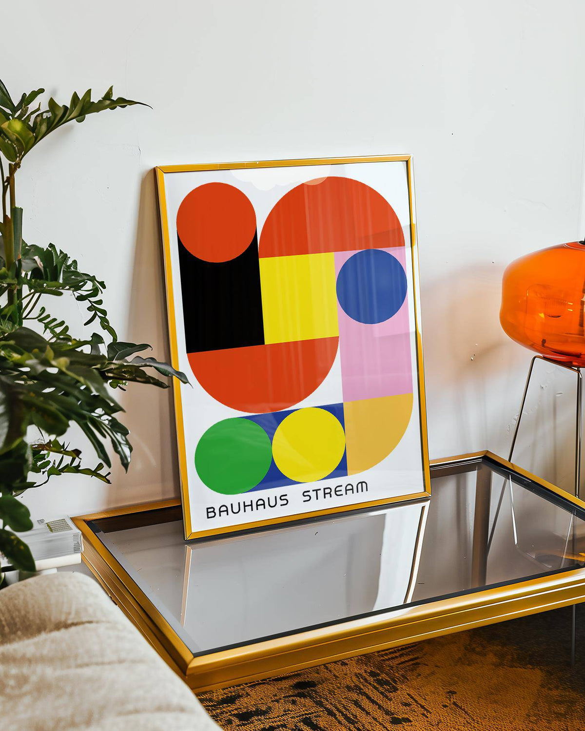 Framed geometric wall decal print showing bold colourful Bauhaus shapes leaning on a glass table next to a plant