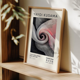 Framed print leaning on a shelf with spiral dot pattern in pink, black and grey with natural light and a plant