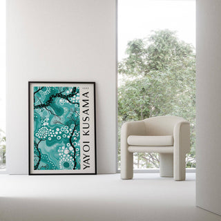 Teal wall decor leaning on floor with abstract branches and dots beside a cream armchair