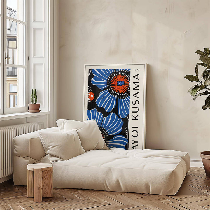 Print leaning on the floor of blue floral artwork with red and black details beside a white sofa and wooden stool