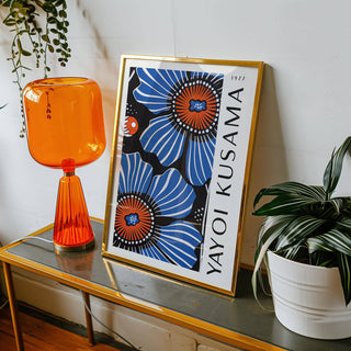 Print leaning on a shelf of blue poppy flowers with orange centres in a gold frame alongside an orange lamp and a plant