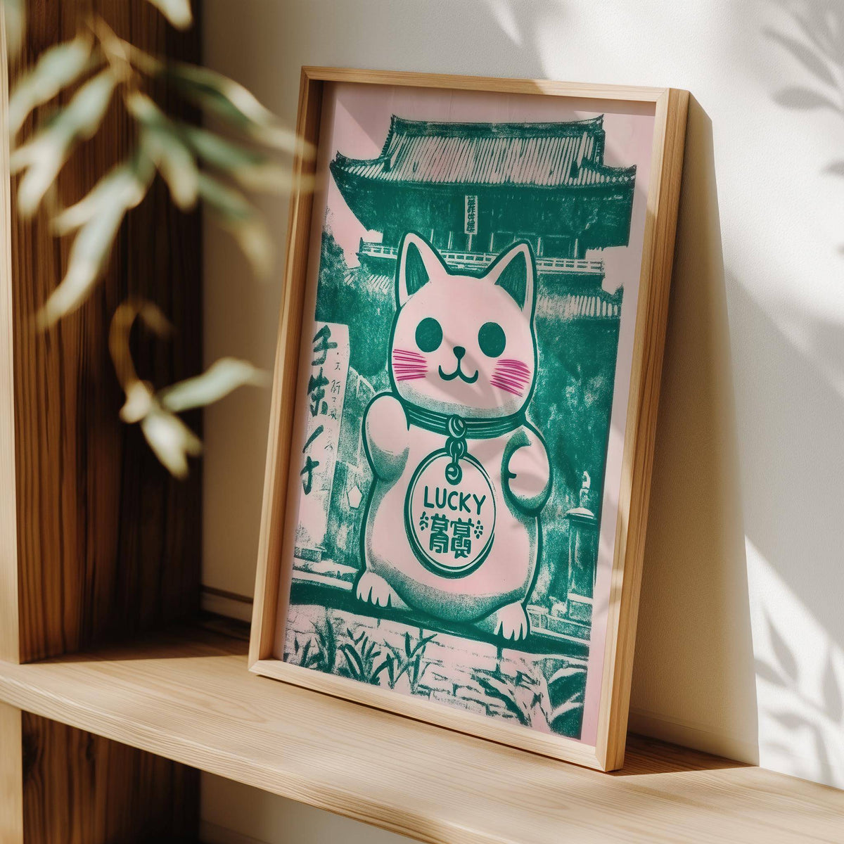Print leaning on a shelf of a maneki-neko cat illustration in green and pink with wooden frame
