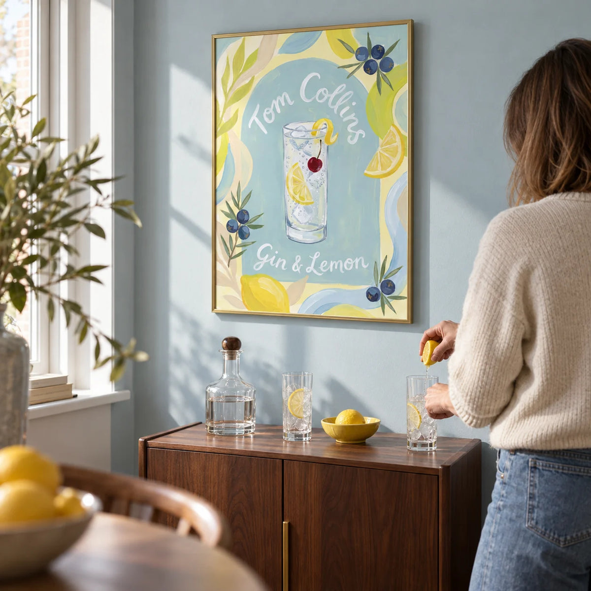 Tom Collins Art Print, framed on the wall of a compact home bar corner in a UK flat with a muted pale blue wall and a slim walnut drinks cabinet in a living room
